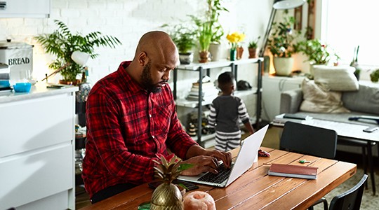 man working from home on a laptop surrounded by house plants with his son playing in the background